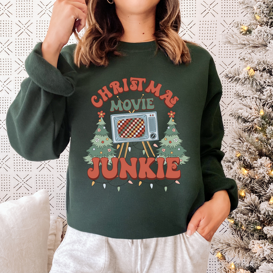 A dark green sweatshirt with the words "Christmas Movie Junkie" surrounding a retro style TV. Two decorated Christmas trees can be found on either side, and Christmas lights hang from the bottom of the design.