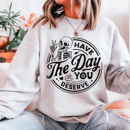 A woman wearing an ash grey sweatshirt with the words "Have the Day you deserve" and a cartoon skeleton giving a peace sign.