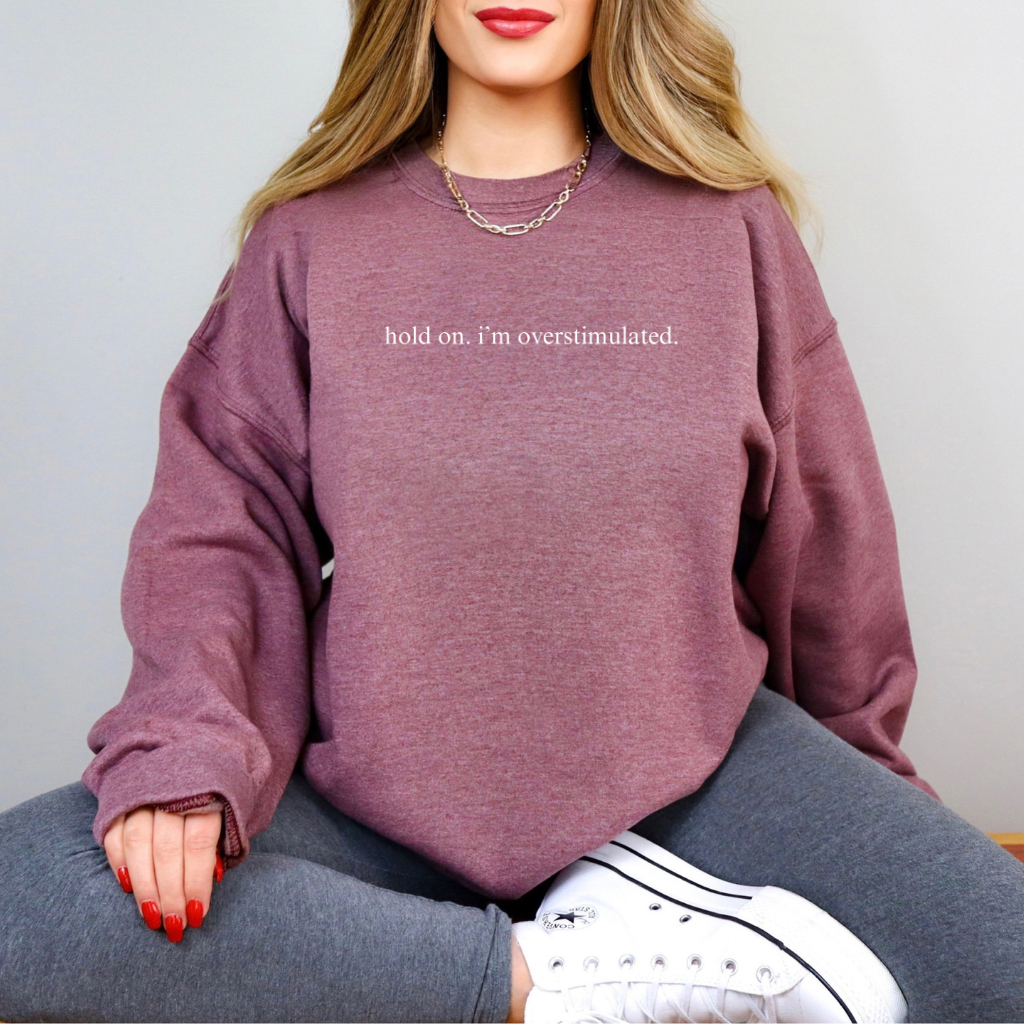 A woman wearing a heather maroon sweatshirt with the words "hold on. I'm overstimulated," written in all lower case.