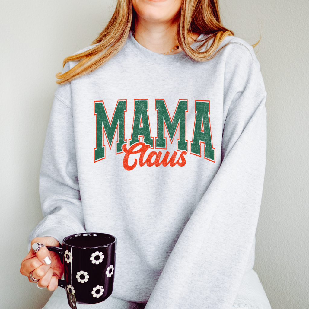 A woman holding a black floral mug wearing a light grey sweatshirt that says "Mama Claus" in green and red retro font.